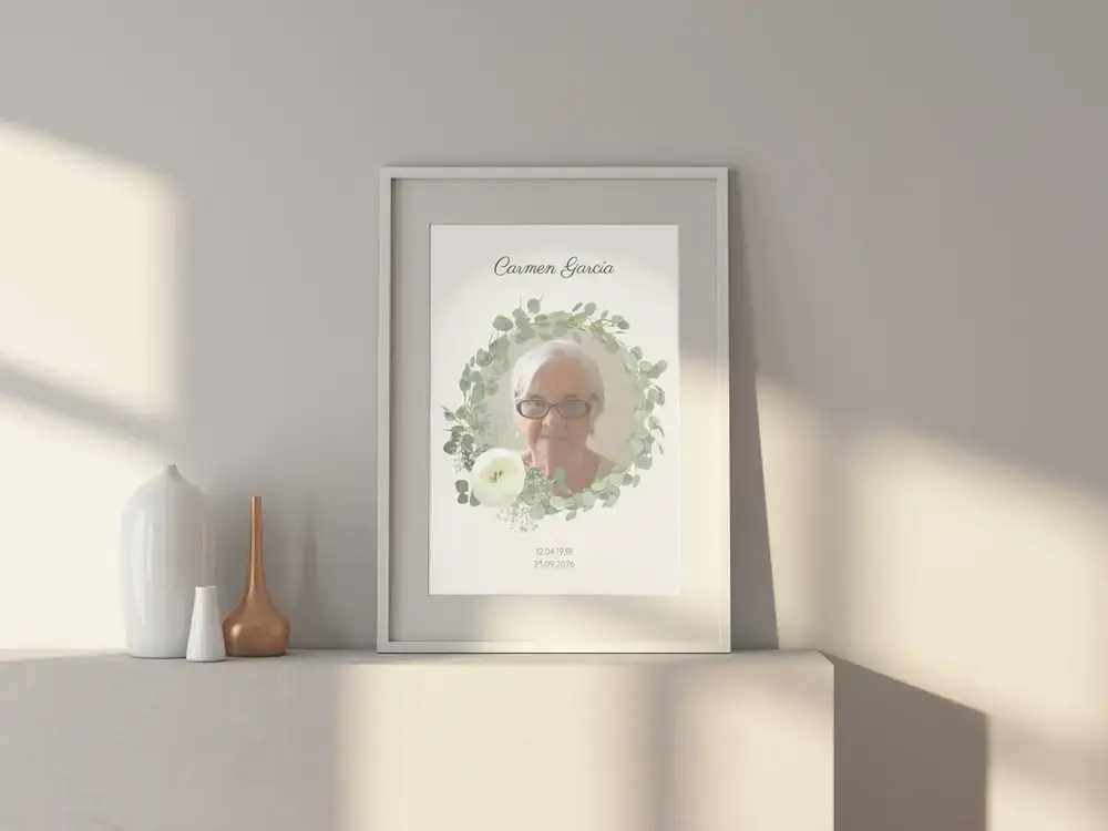 Cuadro conmemorativo con retrato y corona de eucalipto, en marco claro sobre un aparador blanco junto a unos jarrones